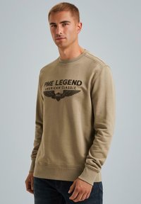 Khaki mikina s okrúhlym výstrihom, s čiernou grafikou "PME LEGEND AMERICAN CLASSIC" a dizajnom krídiel. Mäkký materiál a rebrované manžety.