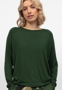 Maglia a maniche lunghe verde scuro con scollo ampio e maniche raglan, realizzata in tessuto morbido con texture liscia. Nessun motivo o accentuazione visibile.