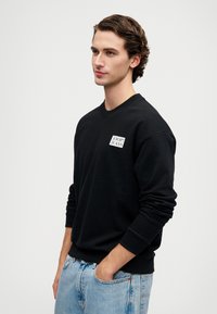 Felpa nera in cotone con polsini e scollatura a coste. Presenta un piccolo logo quadrato sul petto. Abbinata a jeans azzurri chiari.