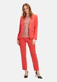 Korálový blazer a kalhoty s košilí s geometrickým vzorem. Hladký materiál, přiléhavý střih, klopové límce a slim kalhoty. Černé špičaté lodičky.