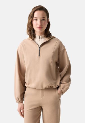 Kvinde med skulderlangt hår iført beige hættetrøje med lynlås og matchende bukser mod ensfarvet baggrund, hænderne i lommerne.
