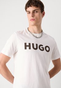 Fehér pamut póló feltűnő fekete "HUGO" felirattal, rövid ujjakkal és kerek nyakkivágással, ezüst lánc nyaklánccal párosítva.