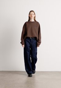 Brown oversized sweatshirt with a crew neck, paired with dark blue wide-leg jeans featuring visible stitching; black shoes complete the outfit.