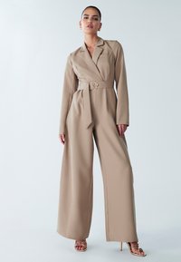 Bēša kopējais tērps ar jostu, garām piedurknēm, plašām kāju biksēm, divām sānu kabatām un izgriezuma apkakli; kombinēts ar zelta krāsas strapatām augstpapēžu kurpēm.