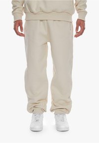 Joggers in cotone beige con polsini elasticizzati e zip laterali. Abbinati a sneakers bianche, caratterizzati da una texture liscia e una vestibilità rilassata.