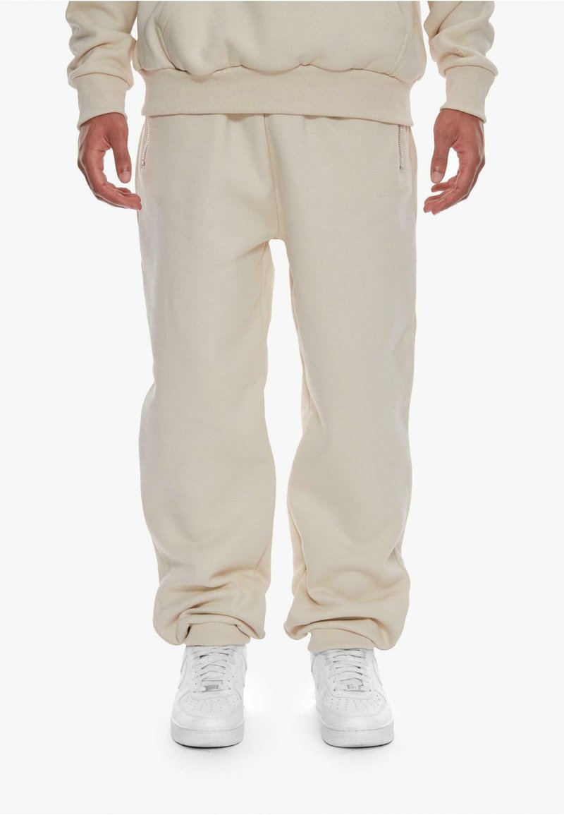 Joggers in cotone beige con polsini elasticizzati e zip laterali. Abbinati a sneakers bianche, caratterizzati da una texture liscia e una vestibilità rilassata.