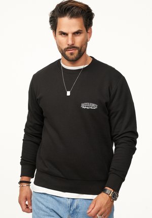 Schwarzer Sweatshirt mit strukturiertem Stoff, Rundhalsausschnitt, gerippten Bündchen und Saum, mit weißem Logo auf der linken Brust. Getragen über einem weißen Hemd.