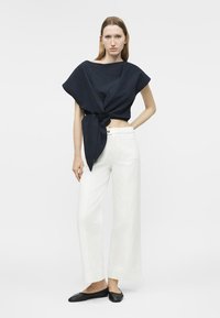 Γυναίκα με αμάνικο, σκουρόχρωμο κορμάκι τύπου wrap δεμένο στη μέση, άσπρο παντελόνι με φαρδιά μπατζάκια και μαύρα επίπεδα παπούτσια, στέκεται με το ένα χέρι στη μέση.