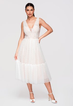 Mujer posando con vestido midi blanco sin mangas con escote en V profundo y falda transparente con lunares, llevando sandalias blancas de tacón alto.