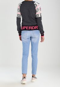 Pull gris avec des manches colorées à motifs, arborant un logo "SUPERDRY" rose sur l'ourlet, associé à un jean en denim bleu clair et des baskets grises.