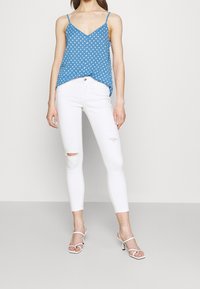 Femme portant un haut sans manches à pois bleu, un jean skinny blanc déchiré et des sandales blanches à talons, se tenant devant un fond blanc uni.