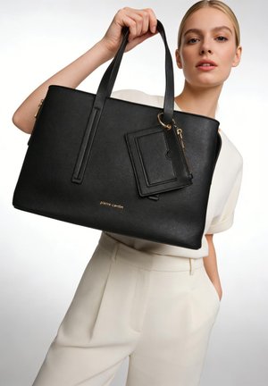 Donna bionda con abito bianco tiene una grande borsa nera Pierre Cardin con portacarte allegato su sfondo bianco.