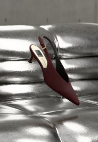 Σκούρο κόκκινο γόβα slingback με τακούνι kitten και μυτερή μύτη, ακουμπισμένη σε μια γυαλιστερή ασημένια επιφάνεια από δερματίνη με κουμπιά.