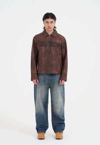 Brun læderjakke med lynlås foran, med sort tekst "Ed Hardy 1971". Kombineret med lyseblå, løstsiddende jeans og brune træsko.