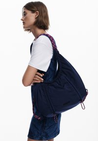 Navyfarbener Stoffbeutel mit einem gerafften Design und einem gemusterten Riemen. Verfügt über ein Logopatch, lebendige Details und eine weiche Textur.