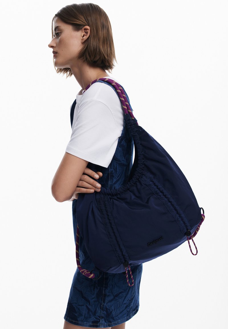 Navyfarbener Stoffbeutel mit einem gerafften Design und einem gemusterten Riemen. Verfügt über ein Logopatch, lebendige Details und eine weiche Textur.
