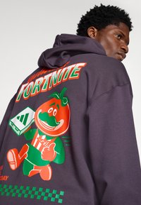 Sweatshirt roxo com um grande personagem de tomate em cartoon, colorido, com um bigode verde, segurando um console de jogos. Nas costas, exibe o texto "Fortnite".