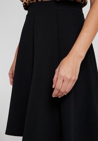 Zwarte rok met een textuurpatroon, halflang, met een aansluitende tailleband en naden. Zichtbare hand die op de stof rust.