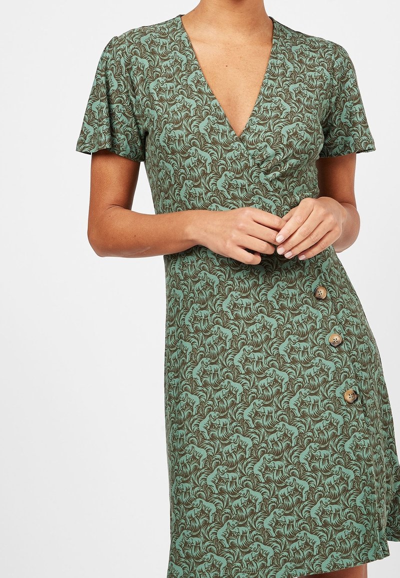 Femme portant une robe verte avec un motif tigre et feuilles, manches courtes, col en V et trois boutons sur un côté près de l'ourlet.