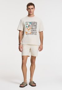 Cream cotton t-shirt with colorful graphic text design, paired with light shorts. Accessorized with a beaded bracelet and green sandals.
