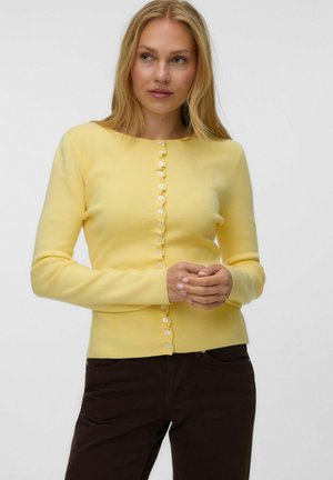 Femme aux longs cheveux blonds portant un cardigan jaune boutonné et un pantalon sombre, debout les mains légèrement croisées, regardant vers sa gauche.
