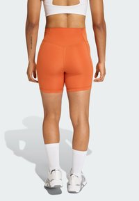 Oranžové šortky s vysokým pásom v priliehavom strihu, vyrobené z elastického materiálu. Majú čisté dokončenie švov a siahajú nad stred stehien.