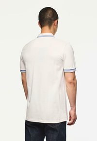 Mies, joka on käännetty poispäin ja pukeutunut valkoiseen polo-paitaan, jossa on siniraitainen kaulus ja hihansuut, yhdistettynä tummansinisiin farkkuihin.