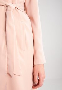 Abito rosa chiaro con cintura annodata in vita, maniche lunghe e due tasche laterali, caratterizzato da una texture liscia e design aderente.