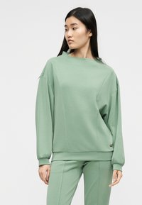 Γυναίκα με μακριά μαύρα μαλλιά, φοράει άνετο φαρδύ φούτερ σε σμαραγδί χρώμα και αντίστοιχο παντελόνι, κοιτάζει προς τα αριστερά της, με φόντο ένα απλό τοίχο.