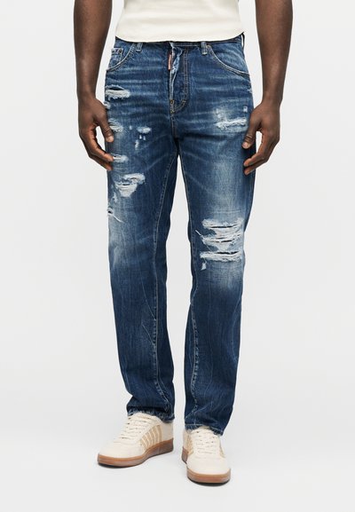 Homme portant un jean bleu déchiré et des baskets blanches, debout devant un fond uni.