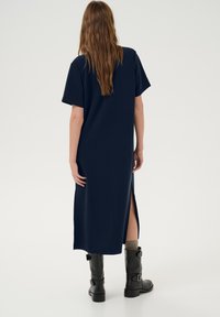 Abito midi blu navy con maniche corte, taglio dritto, spacchi laterali e texture liscia; indossato con stivali neri fino al ginocchio.