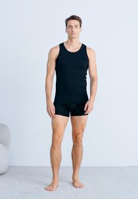 Un débardeur noir et un short de boxeur assorti portés par un mannequin masculin debout sur un fond clair, mettant en valeur un design lisse et ajusté.