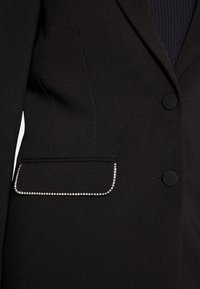 Blazer noir sur mesure avec deux boutons recouverts de tissu et un rabat de poche orné de petits strass étincelants.