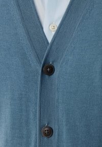 Cardigan tricoté bleu avec deux boutons noirs sur une chemise bleu clair avec un bouton crème visible.