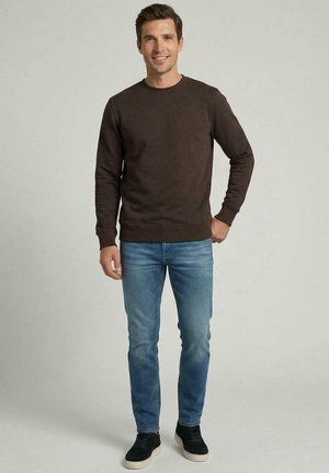 Glunderende man met een donkerbruine trui, blauwe spijkerbroek en zwarte sneakers, staande met één hand in zijn zak tegen een effen achtergrond.