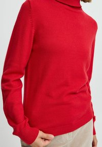 Maglione a collo alto lavorato a maglia rossa con polsini e orlo a coste. Texture morbida, design aderente. Il modello indossa pantaloni in corduroy beige.