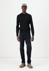 Maglione nero a maniche lunghe con scollo a V, jeans blu scuro e scarpe robuste marrone chiaro. Un cappellino grigio chiaro con del testo poggia sulla testa.