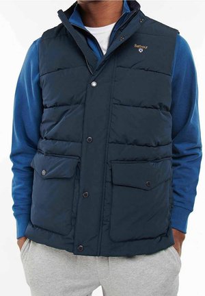 Homme portant un gilet matelassé Barbour bleu marine sur une chemise bleue à manches longues, les mains dans les poches, vu du cou aux hanches.