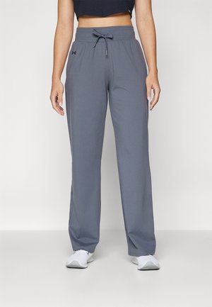 Kobieta ubrana w szare spodnie dresowe ze sznurkiem, czarny krótki top i białe tenisówki, stojąca na tle jasnego, jednolitego tła.