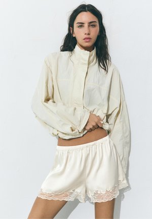 Kvinde med langt mørkt hår iført en cremefarvet højhalset jakke og satin cremefarvede shorts med blonderkant, stående mod en ensartet hvid baggrund.