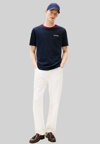 Maglietta blu navy con rifiniture rosse e bianche, pantaloni bianchi e mocassini scuri. Presenta un logo ricamato sul petto. Design semplice e casual.