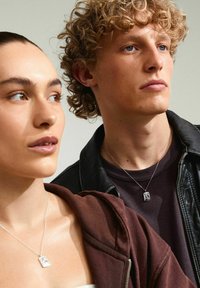 Colgantes de plata en cadenas finas llevados por dos modelos, uno con una sudadera marrón oscuro y el otro con una chaqueta de cuero negra, fondo claro.