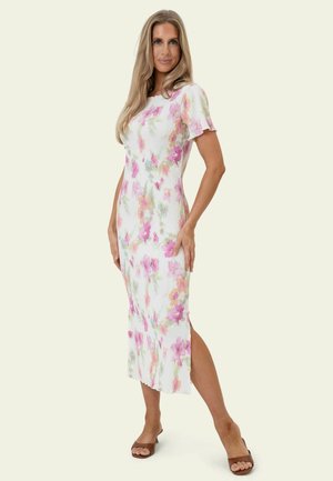 Kvinde stående med den ene hånd på hoften iført en hvid midi kjole med pink og orange blomsterprint og brune åbne hæle.