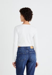 Biała długorękawowa crop top noszona z wysokim stanem ciemnoniebieskimi dżinsami, z skórzaną łatą na pasku i tylnych kieszeniach.