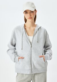 Felpa grigia chiara con zip e cappuccio, dotata di stringhe, caratterizzata da due tasche frontali e polsini a coste, indossata sopra una maglietta bianca.