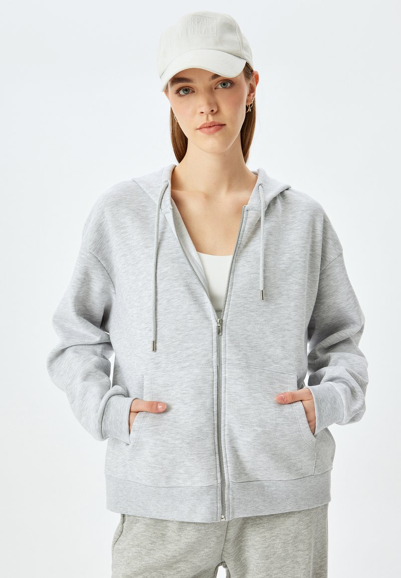 Felpa grigia chiara con zip e cappuccio, dotata di stringhe, caratterizzata da due tasche frontali e polsini a coste, indossata sopra una maglietta bianca.