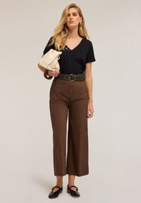 Mujer vestida con camiseta negra de cuello en V, pantalones de cintura alta marrones de pierna ancha con cinturón, zapatos planos negros, sosteniendo un bolso beige sobre un fondo neutro.