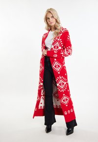 Roter Strickcardigan mit geometrischem weißen Muster, langen Ärmeln, offener Front und weicher Textur, kombiniert mit schwarzen Schlaghosen.
