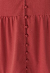 Punane kangas tekstuurse viimistlusega, mille keskel on rida kuus ümarat nööpi ja vööjoonel kogutud detailid.