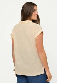 Camicia corta a maniche corte color beige chiaro con design in rete e colletto solido. Presenta una vestibilità comoda e un orlo che arriva appena sotto la vita.